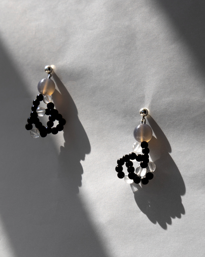 A pair of statement dangle earrings with black onyx beaded loops, clear round grey agate beads, and silver-toned posts, displayed on a textured light gray surface with dramatic shadows.