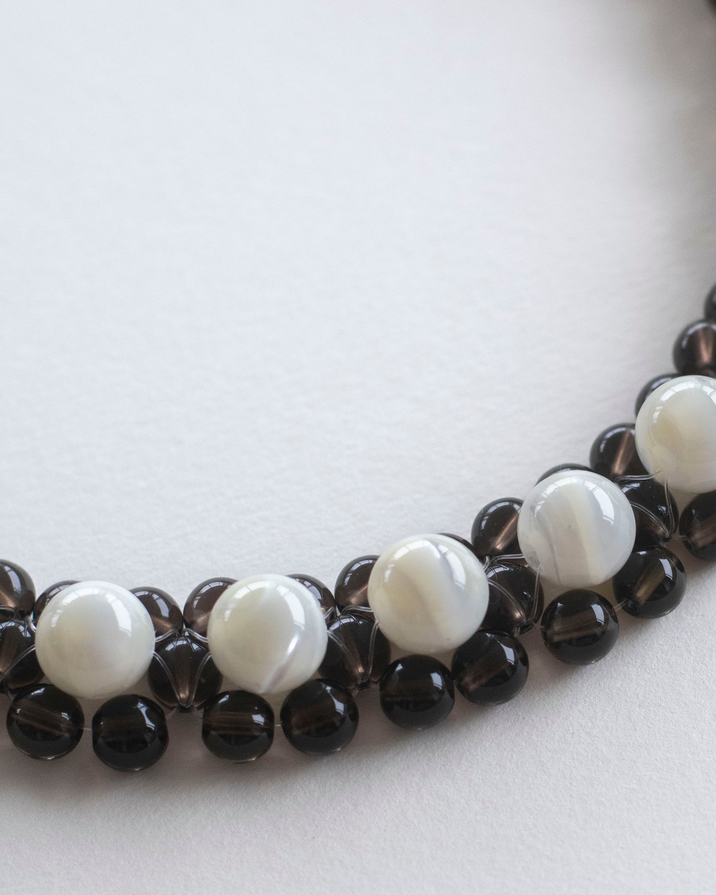 A close-up of a beaded necklace choker featuring alternating smooth white mother of pearl beads and dark brown smoky quartz beads, arranged in a curve on a plain light background.