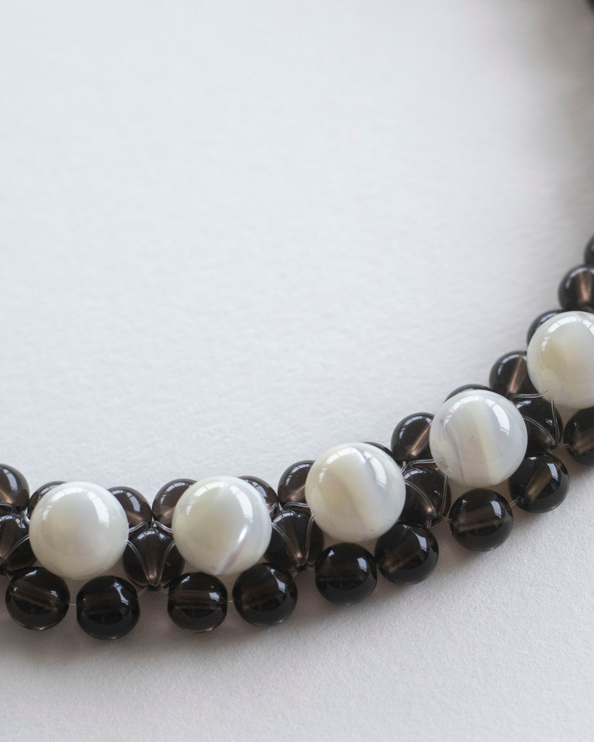 A close-up of a beaded necklace choker featuring alternating smooth white mother of pearl beads and dark brown smoky quartz beads, arranged in a curve on a plain light background.