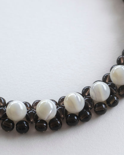 A close-up of a beaded necklace choker featuring alternating smooth white mother of pearl beads and dark brown smoky quartz beads, arranged in a curve on a plain light background.