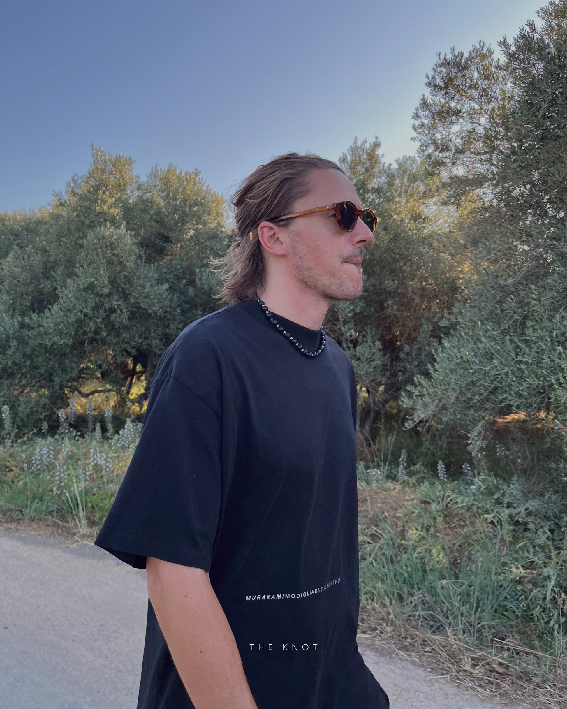 Man wearing TERRA006 black matte agate beaded necklace, with sunglasses and a black t-shirt with text, standing outdoors with trees in the background