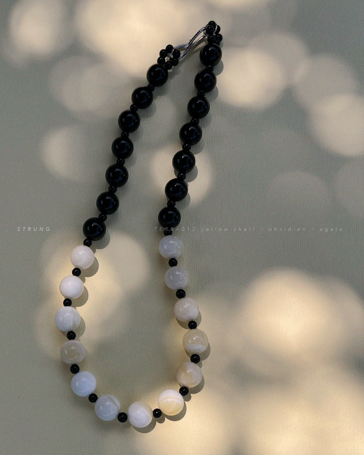 TERRA012 necklace featuring handstrung warm yellow shell beads, black obsidian, and black agate on silk thread with rhodium-plated brass clasp witj soft shadows background.