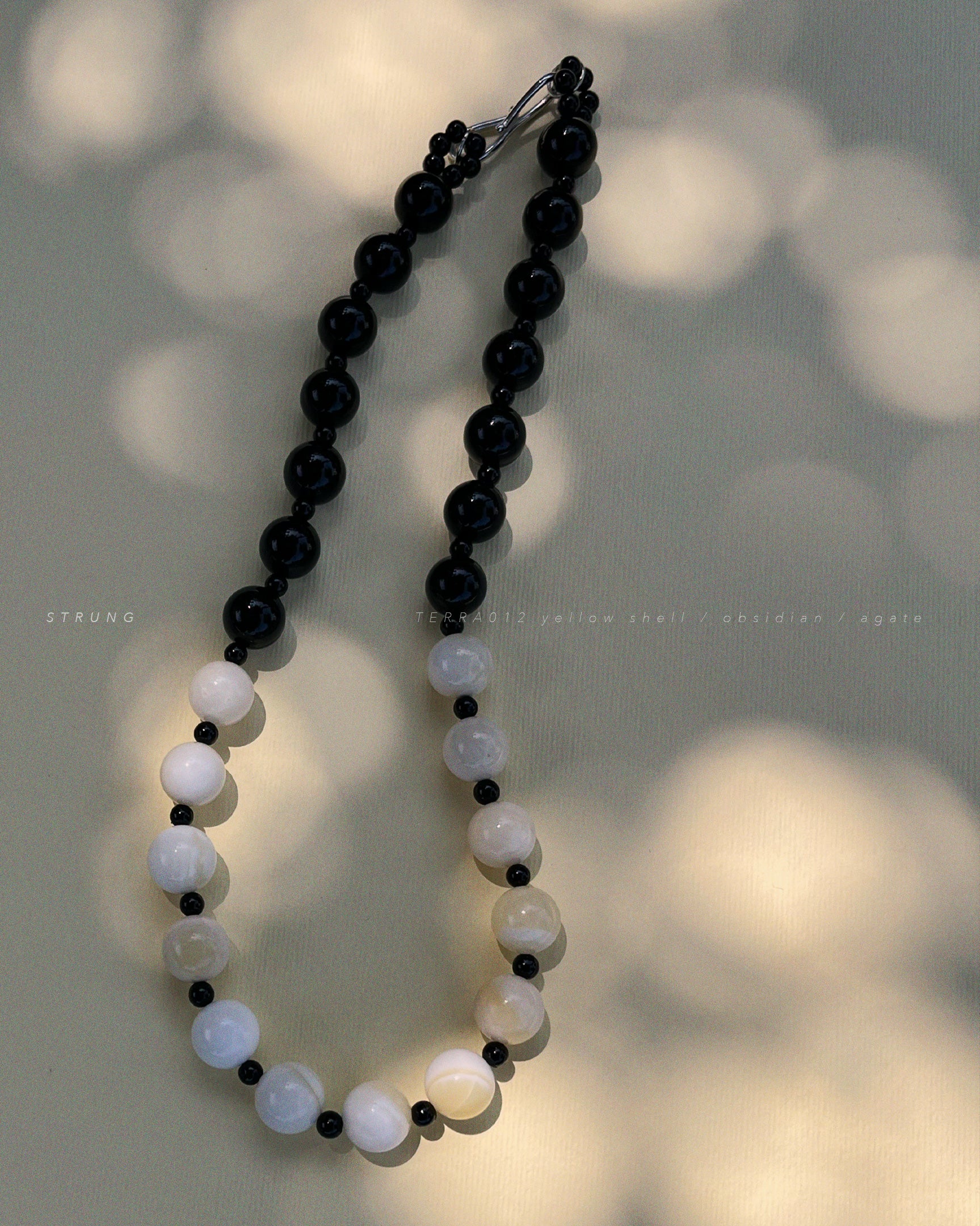 TERRA012 necklace featuring handstrung warm yellow shell beads, black obsidian, and black agate on silk thread with rhodium-plated brass clasp witj soft shadows background.