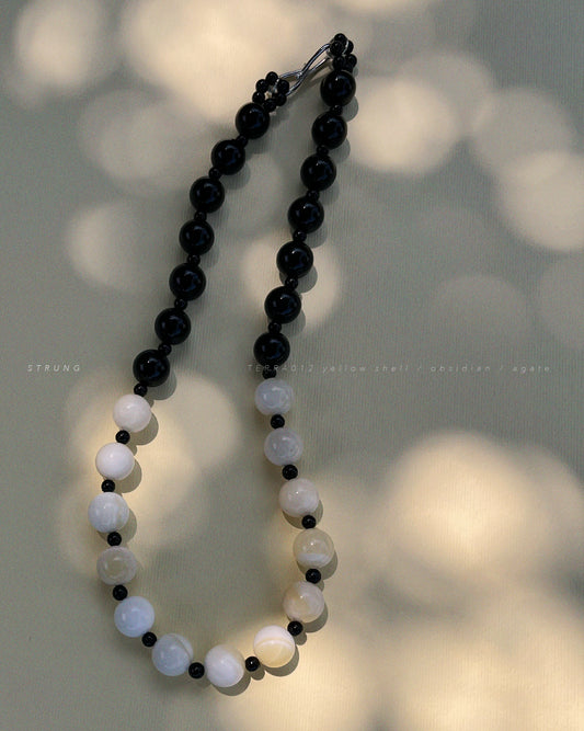 TERRA012 necklace featuring handstrung warm yellow shell beads, black obsidian, and black agate on silk thread with rhodium-plated brass clasp witj soft shadows background.