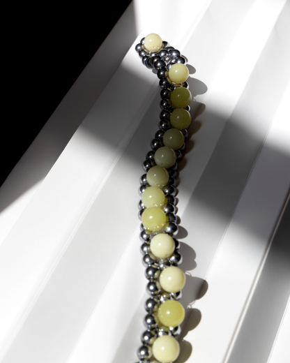 A beaded NECKLACE featuring alternating layers of lemon jasper stones and silver hematite, styled on a white geometric surface with dramatic shadows.